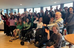 Photo of lots of people (attending Richmond AID's Business Plan Meeting) - some standing, some seated in wheelchairs all waving.