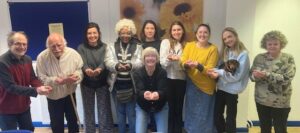 Photo of a group of people who are some of the RAID (Richmond AID) team, clients and volunteers. All standing with outstretched arms and cupped hands demonstrating the theme for 2026 International Women's Day - When we give, we gain!
